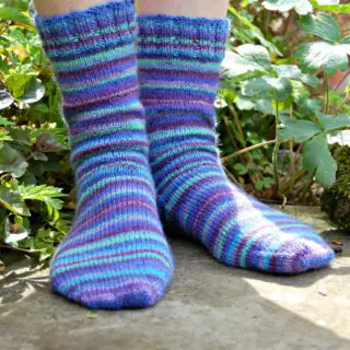 Close-up of feet wearing knitted socks with blue, green, and purple stripes, standing on a stone surface surrounded by leafy greenery. Winwick Mum | Basic 4ply Socks | Beginner sock knitting | Free sock knitting patterns