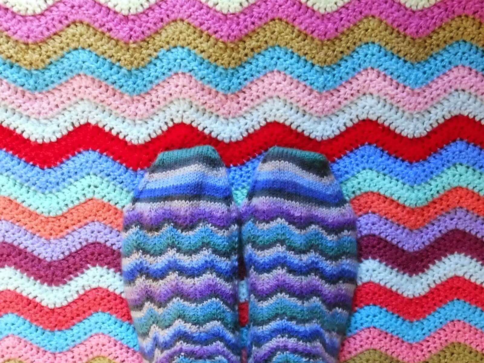 A close up of rippled pattern socks in stripes of blue, grey and green on a background of a colourful rippled crochet blanket