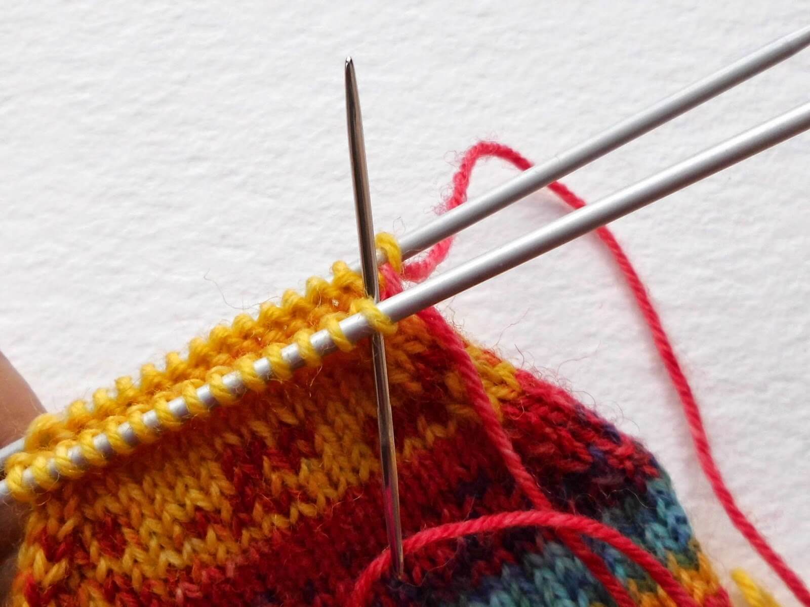 Close-up of two knitting needles with vibrant yarn in red, yellow, and orange hues. A wool needle threads through stitches to close the toe seam conveying a focused crafting process. Winwick Mum Sockalong
