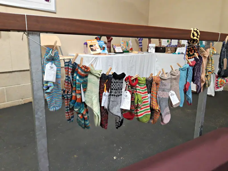 A photograph showing seventeen pairs of socks which are pegged to the Yarndale Sock Line. They are hanging down as if on a washing line. In the foreground is a padded maroon bench seat and in the background is the Winwick Mum stand; two tables covered with white table cloths and displaying Winwick Mum books, socks and yarn