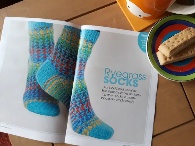 The main image of the photo is a magazine pattern picture of a pair of socks knitted in WYS Winwick Mum yarn Brightside and WYS Signature yarn Bubblegum in a mosaic pattern. There is an orange Herdy mug and a saucer with two shortbread biscuits to the right of the picture