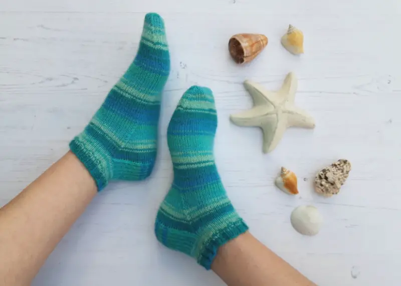 Two feet wearing cosy, teal and blue striped socks rest on a light wooden floor. Nearby are seashells and a starfish, evoking a calm, beachy vibe. Winwick Mum | Basic 4ply Shortie Socks | Beginner sock knitting | Free sock knitting patterns