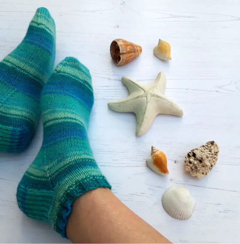 Two feet to the left of the photo wearing cosy, teal and blue striped socks rest on a light wooden floor. Nearby are seashells and a starfish, evoking a calm, beachy vibe. Winwick Mum | Basic 4ply Shortie Socks