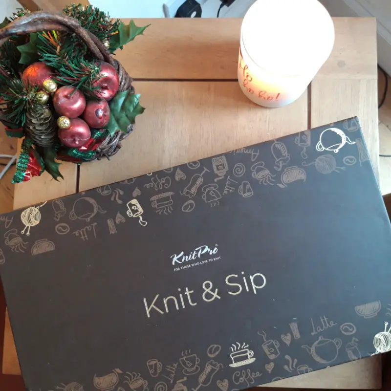 A "Knit & Sip" kit box lies on a wooden table, adorned with playful knitting and coffee-themed illustrations. Beside it, a festive arrangement with red apples and pinecones, and a glowing candle create a cozy, holiday ambiance.