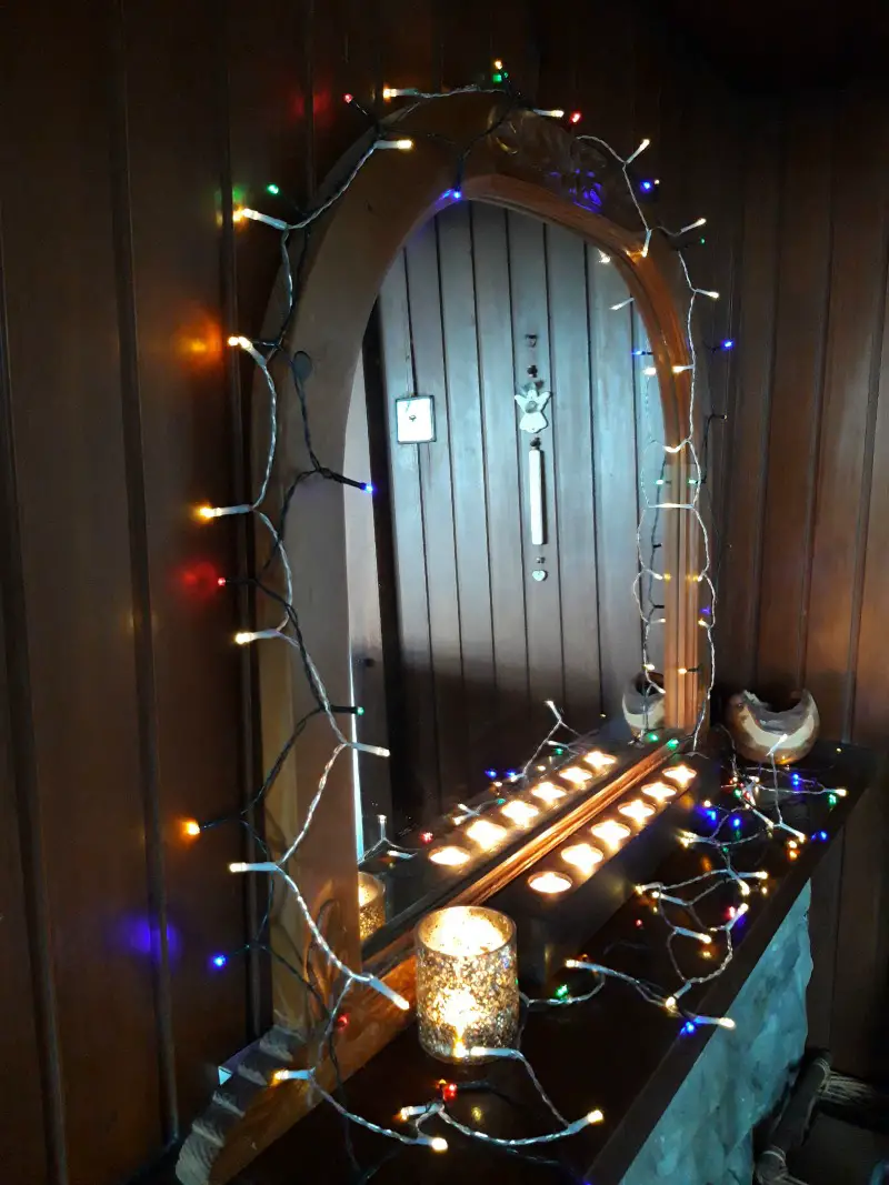 A photo of a mirror in curved wooden frame decorated with multi coloured fairy lights and with candles glowing next to it