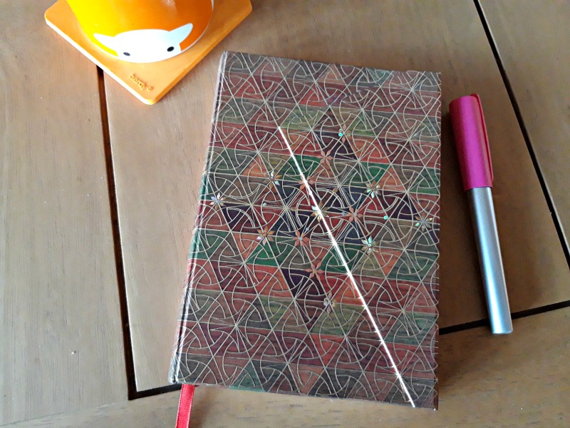 A photo of a pale oak wooden table with an orange mug, a fountain pen and notebook with an ornate design on it