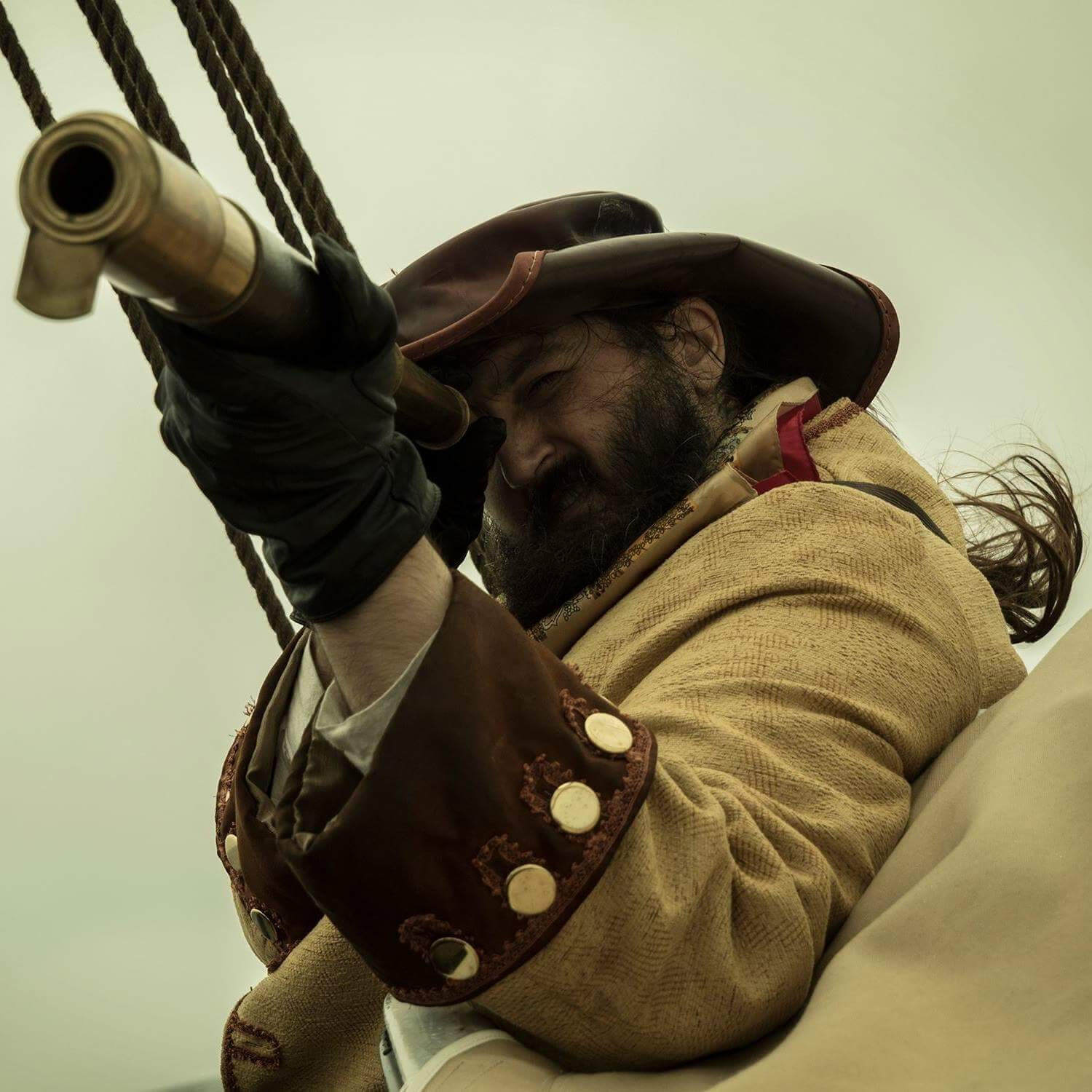 A bearded man is dressed as a pirate in a beige coat and brown hat. He is holding a gun and looking down the sights, standing on board a pirate ship