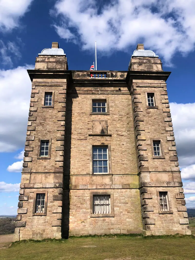 A square stone building on a grassy hill. There are 8 windows and turrets on each side.