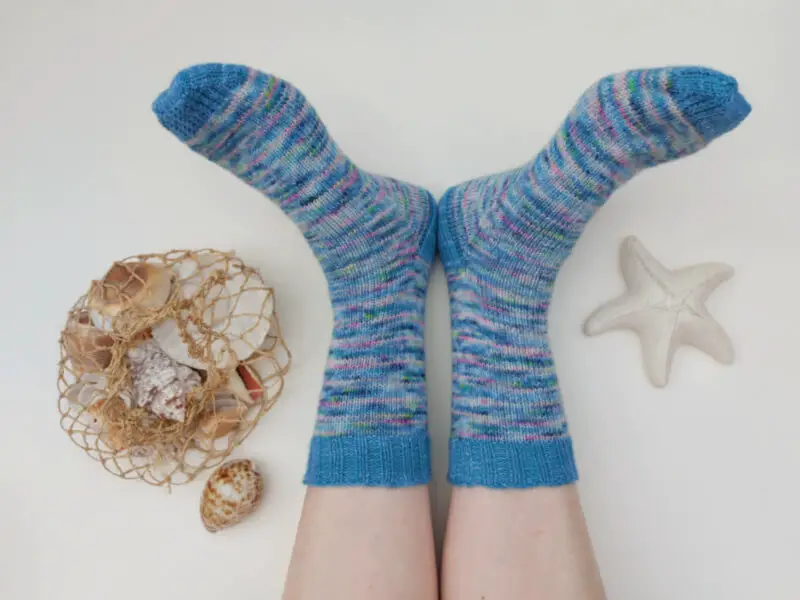 A pair of blue variegated socks with solid blue cuffs, heels and toes are modelled to look like a mermaid's tail. There is a bag of shells to the left and a ceramic starfish to the right of the socks.
