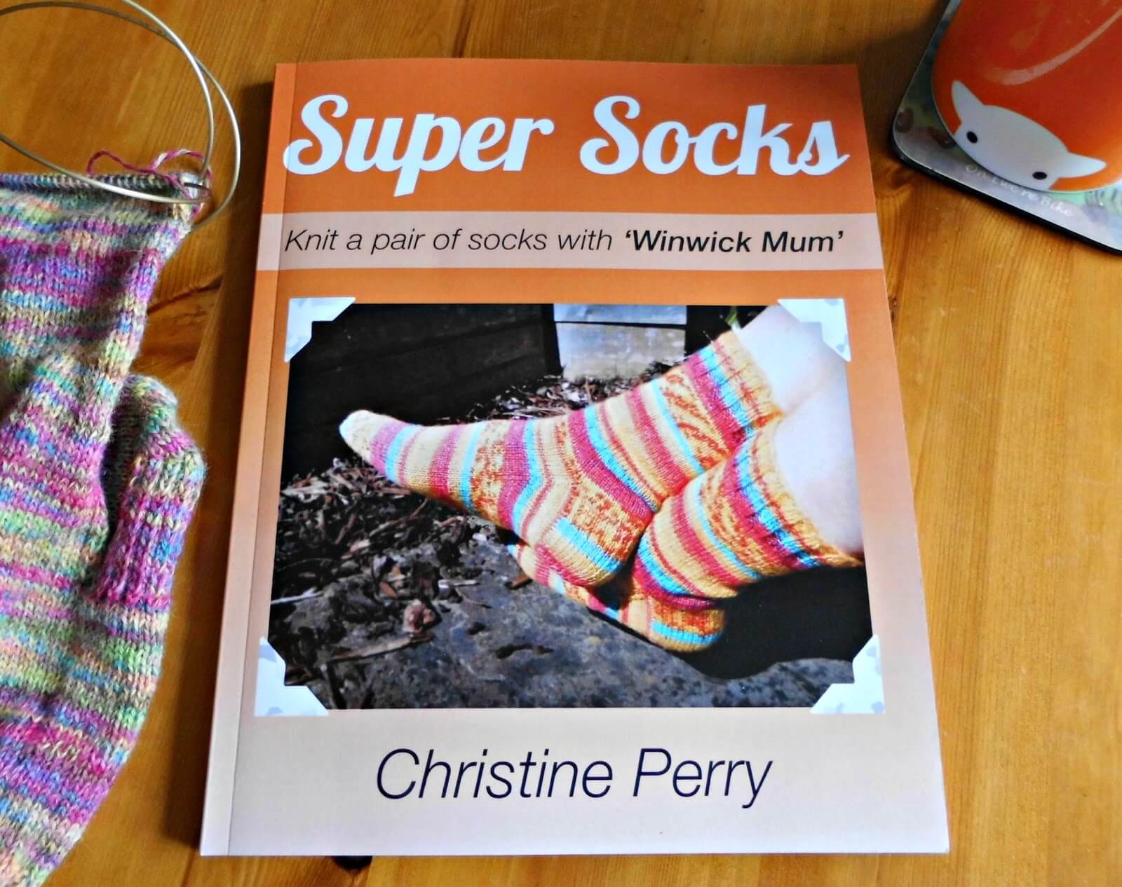 An orange Super Socks book lying on a wooden table. To the left is a partly knitted sock, to the right is an orange mug