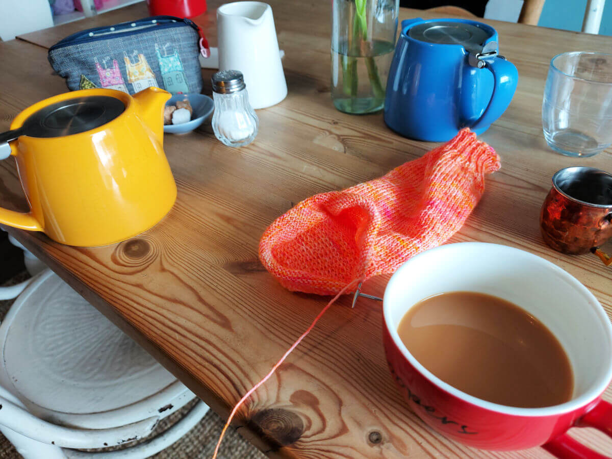 Coloured tea pots, cups, a sugar bowl and salt cellar on a wooden table. There is also a partly-knitted orange sock and a small blue accessories bag with houses on it