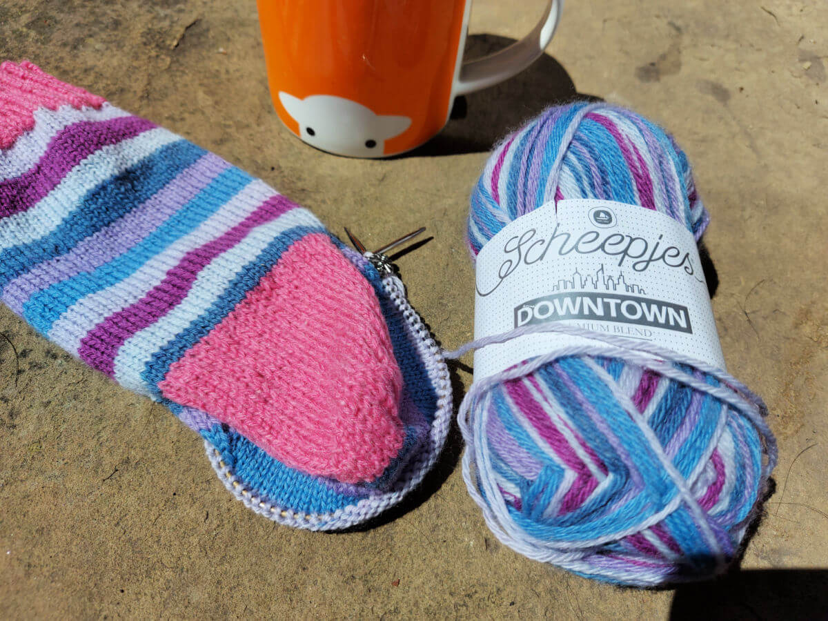 A half-knitted blue, purple and grey stripy sock with a pink heel rests on a stone flag next to a ball of the yarn. Behind is an orange mug of tea.