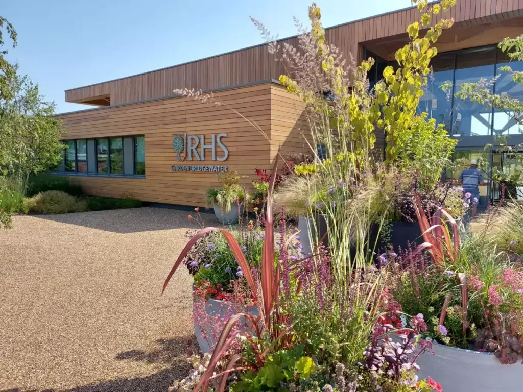 A selection of huge planters filled with plants in front of the RHS Bridgewater welcome building. The wooden-clad centre is behind with the RHS name and logo in silver letters
