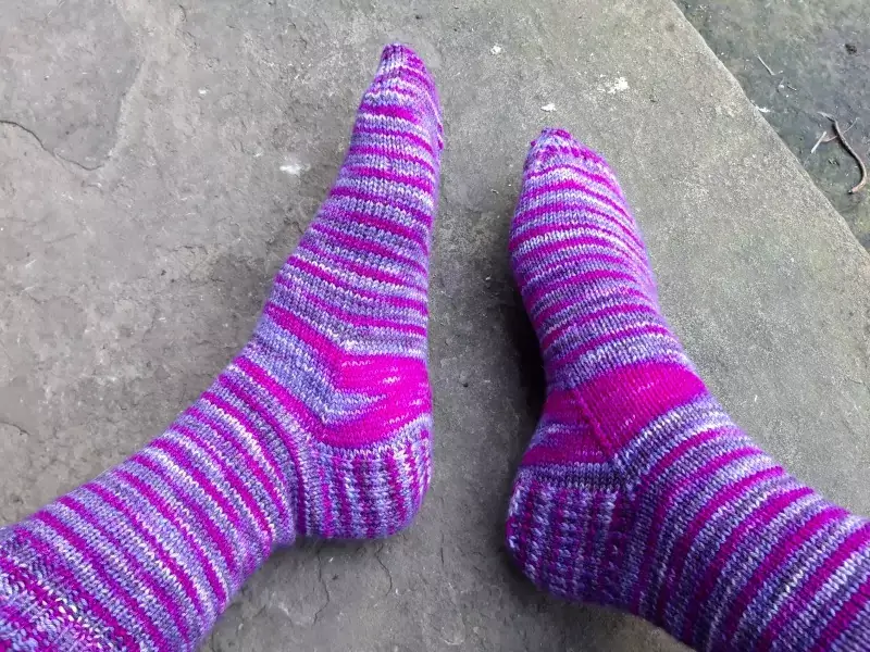 A pair of purple variegated socks modelled on feet, resting on a stone slab. There is a bright pink flash of colour across the gusset of both socks