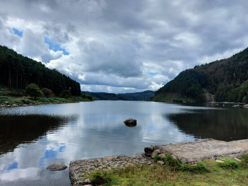 A large lake stretching into the distance, bordered by hills covered in trees. The cloudy sky is reflected in the water.