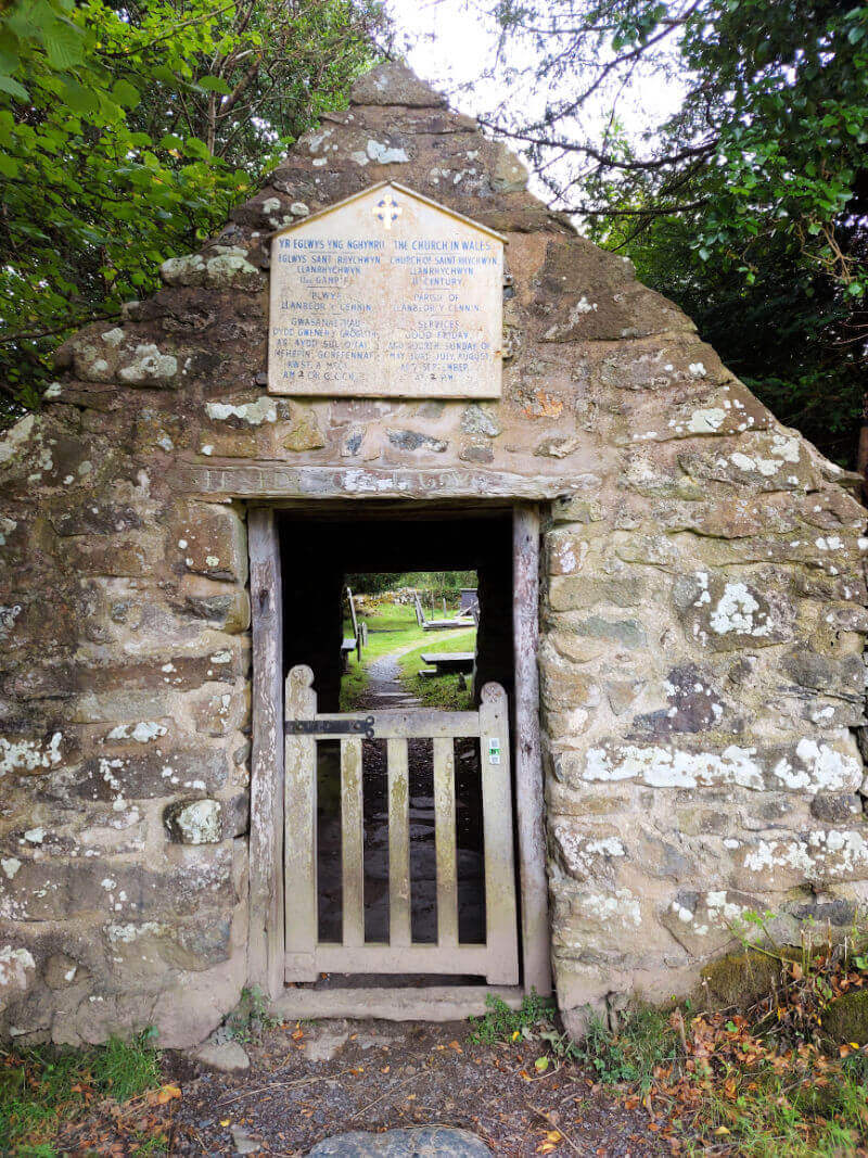 An old stone wall with a gate leading into a church yard. There is a plaque written in Welsh and English above the gate. 
