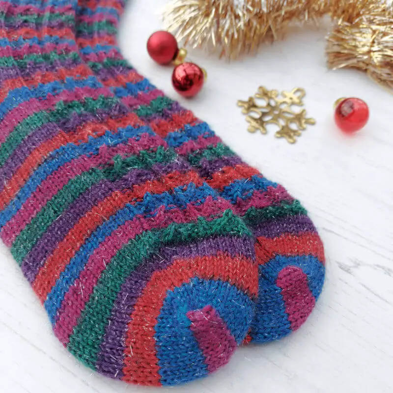 Close up of the Twinkle Toes socks to show the colours at the toes.  The socks are lying on a white wooden board.  At the top of the photo are small red glass baubles, a brass snowflake tree ornament and gold tinsel