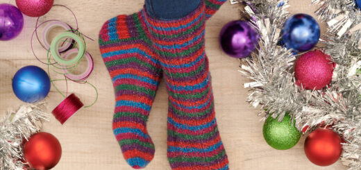 A pair of green, blue, pink, red and purple sparkly socks modelled on feet. The model is surrounded by tinsel, baubles and rolls of gift ribbon.