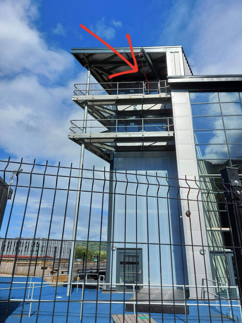 An tall building behind a wire safety fence. There is a red arrow pointing to a metal gate at the top of the building and crash mats at the bottom.