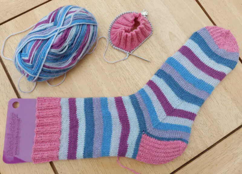 A blue, beige, cream and maroon sock with pink cuff, heel and toes lies on a wooden table. Above it are the pink cuff of the second sock on the circular needle and the ball of striped yarn.