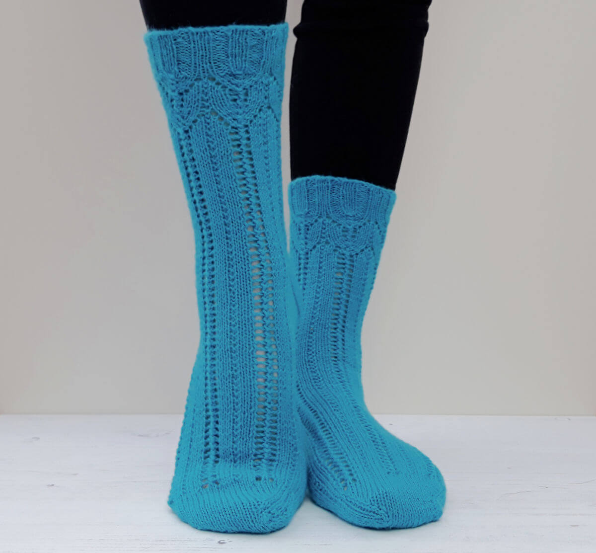 A pair of blue lace patterned socks modelled against a cream background. One heel is slightly raised.