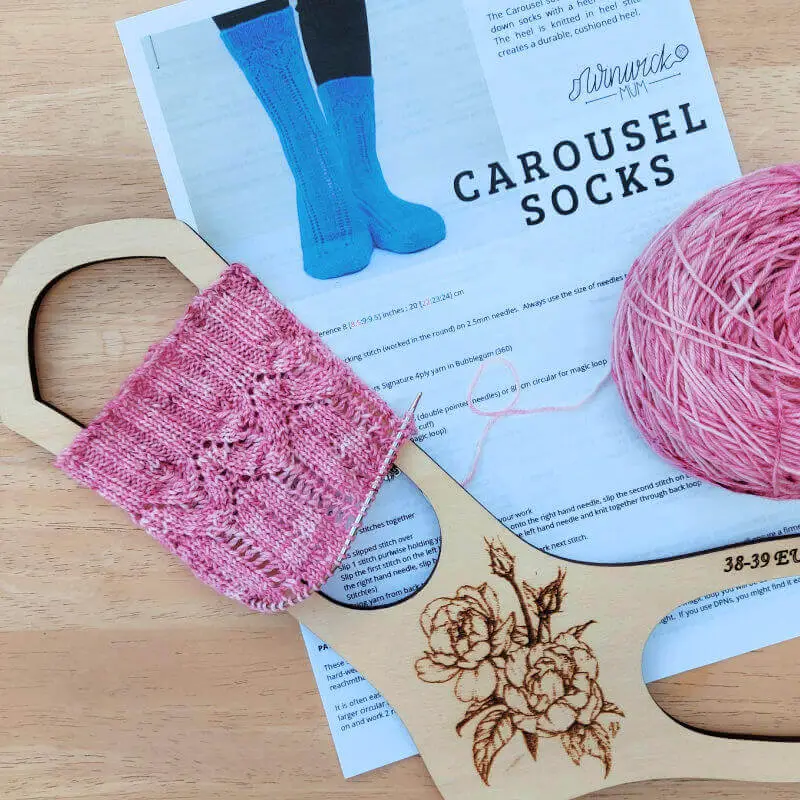 A partly knitted sock is stretched across a wooden sock blocker. The sock is lying on a paper knitting pattern and next to it is a wound cake of pink yarn. Everything is on a wooden table.