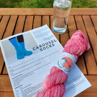 A skein of pink yarn lies across a knitting pattern on a wooden garden table next to a glass of lemonade