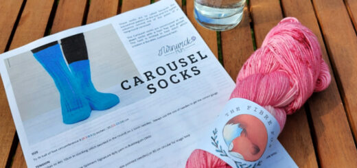 A skein of pink yarn lies across a knitting pattern on a wooden garden table next to a glass of lemonade