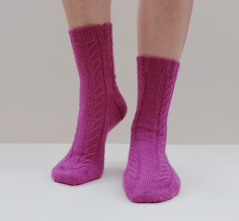 A pair of pink socks with a twisted stitch pattern modelled against a cream background. One heel is slightly raised.