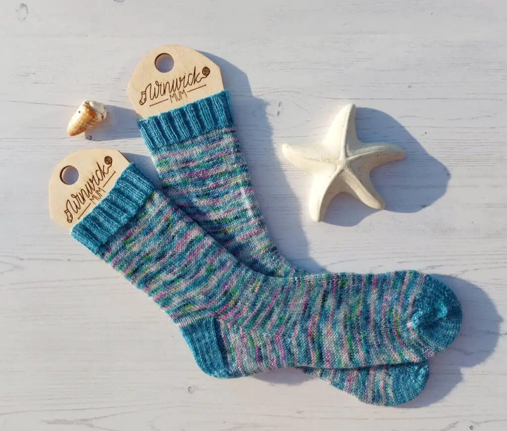 A pair of blue variegated socks on wooden sock blockers against a white wooden board background. There is a shell and a ceramic starfish next to the socks.