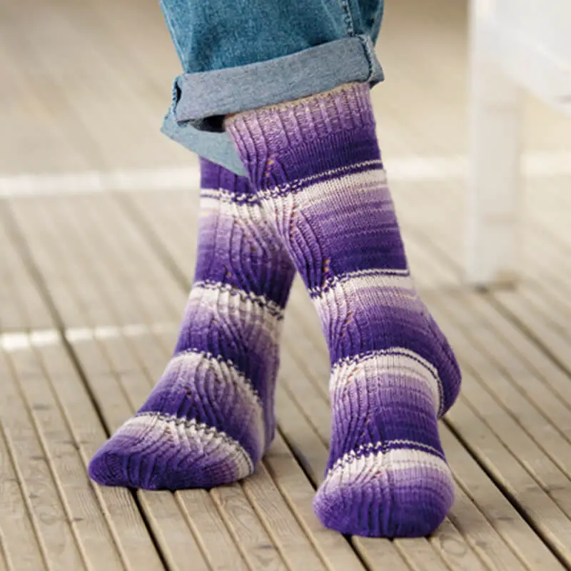 A pair of purple and white striped socks with a twisted stitch panel pattern modelled on feet. One foot is crossed over the other, the toes touching the wooden board floor.