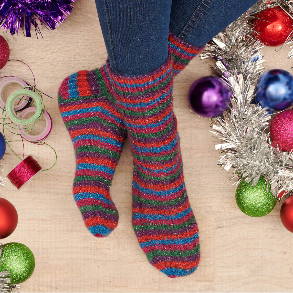 A pair of red, blue, purple, green and pink striped sparkly socks modelled on feet. The feet are crossed at the ankle and surrounded by Christmas tinsel, baubles and gift ribbon.