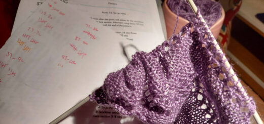 A partly-knitted purple shawl on two long double pointed needles. A sheet marked with rows worked and the pattern are visible, and one of the needles is pushed into a knitting belt