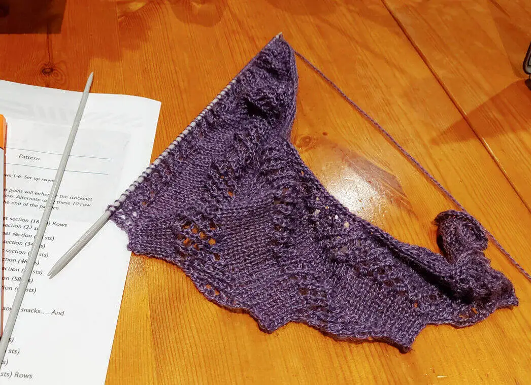 A half-knitted shawl lies on a wooden table on top of the pattern.