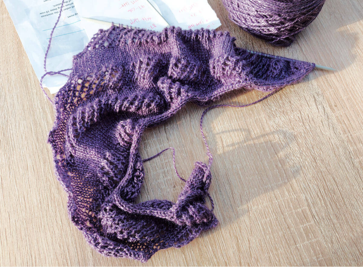 The same half-knitted shawl lies on a different table. Also in the photo this time is a purple ceramic yarn bowl.