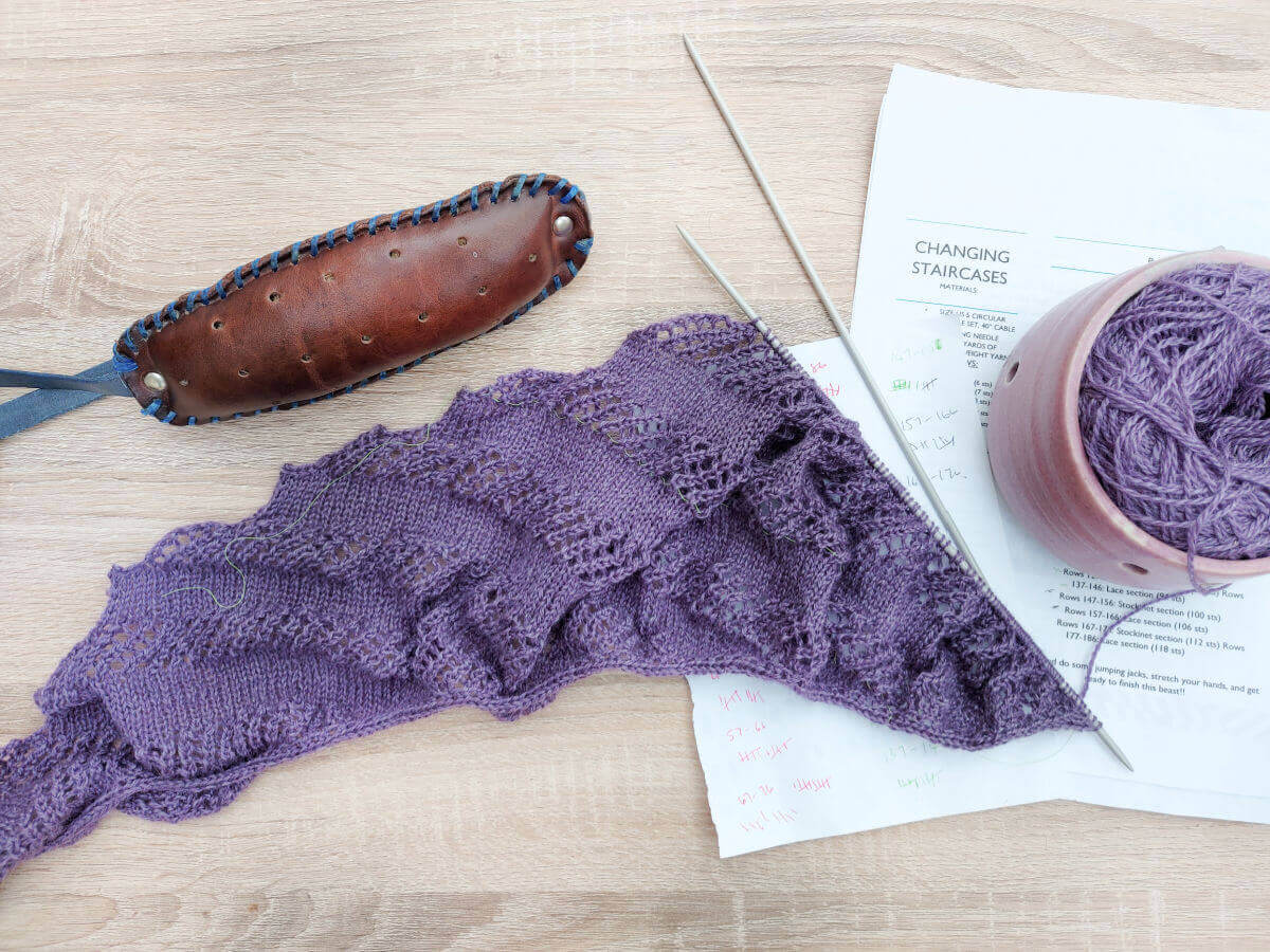 A purple shawl on a knitting needle.  Above the shawl is a brown leather knitting belt and to the right is a pink ceramic yarn bowl containing a cake of purple yarn sitting on the knitting pattern.
