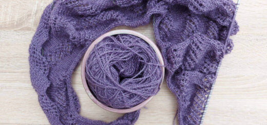 A flatlay of a half-knitted purple shawl curled around a yarn bowl containing the rest of the yarn.