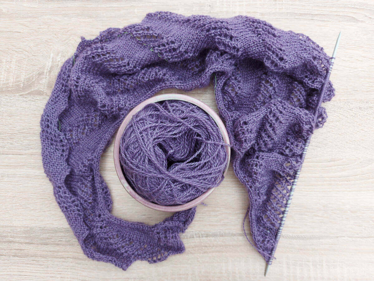 A flatlay of a half-knitted purple shawl curled around a yarn bowl containing the rest of the yarn.