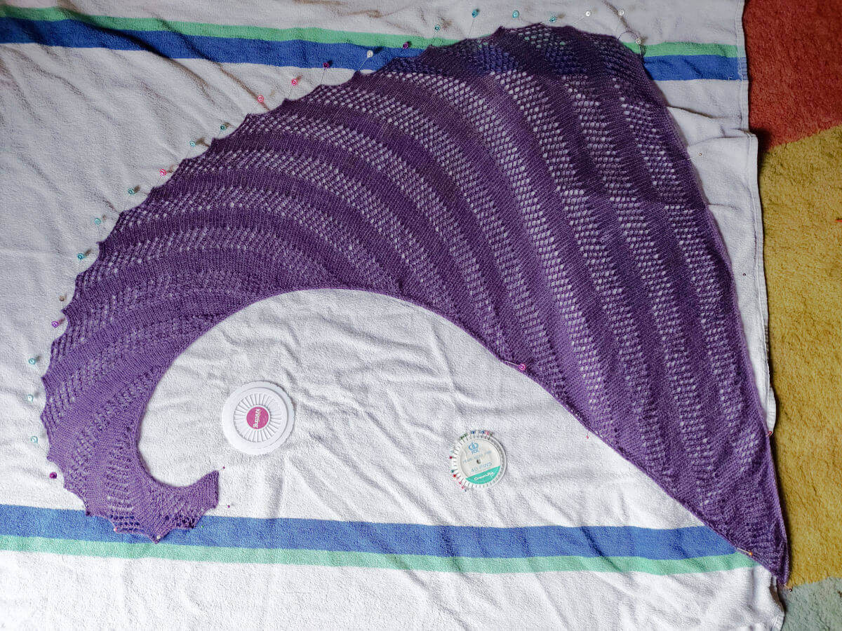 A purple shawl with rows of knit and a lattice stitch is pinned out on a blue and white sports towel.
