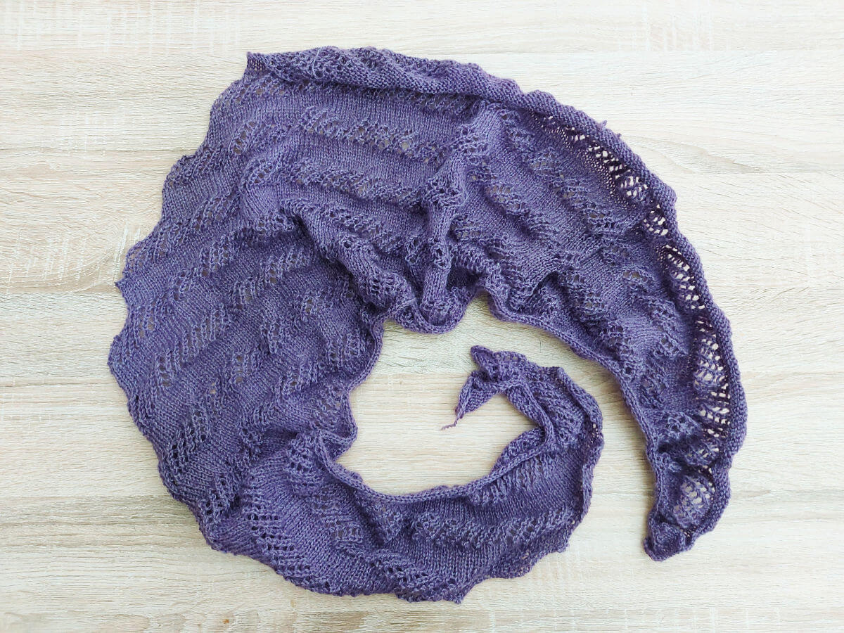 A purple shawl curled around itself on a wooden table. The shawl needs blocking to stretch it out.