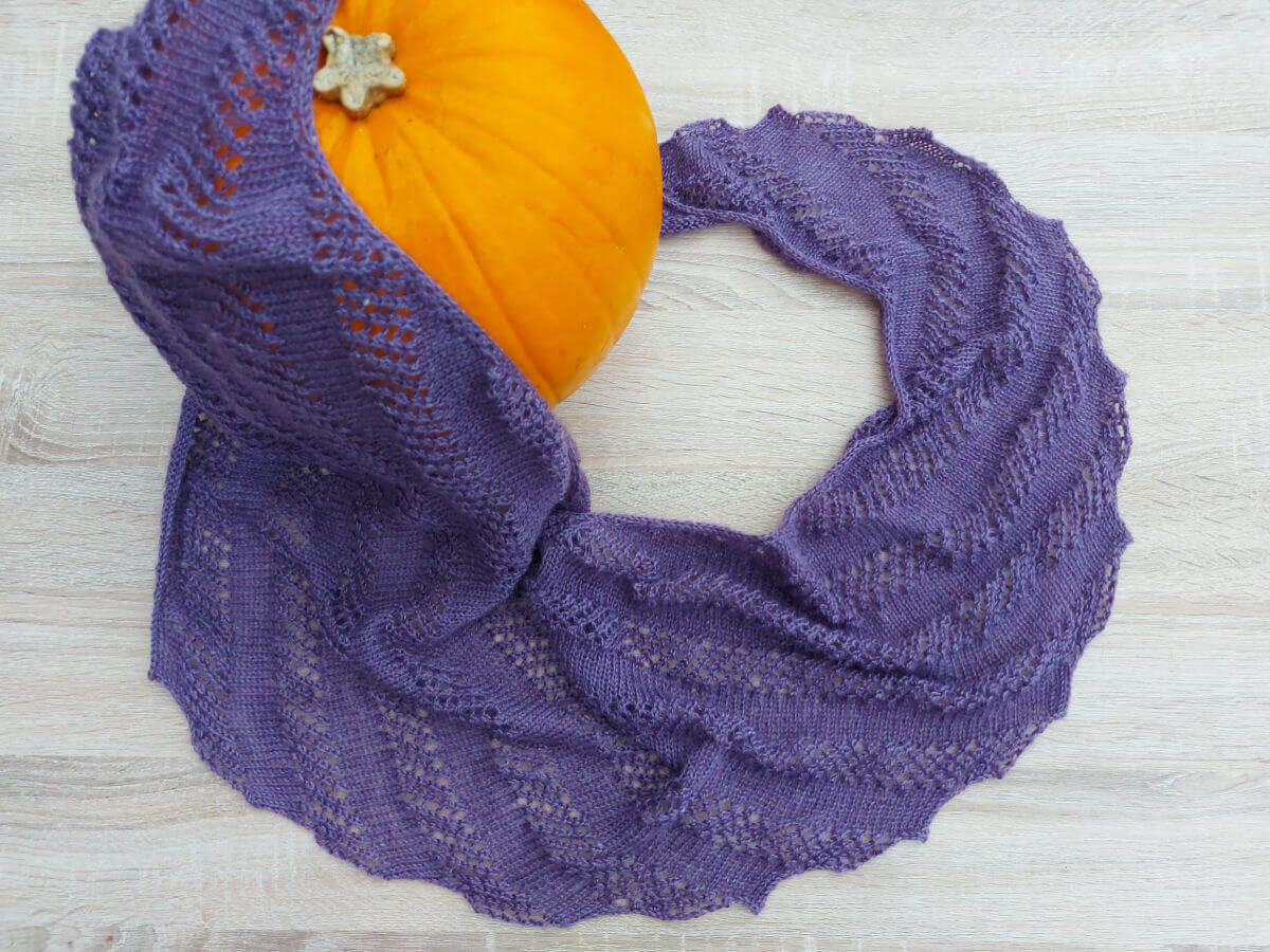 A purple shawl is arranged on a wooden table. One end of the shawl is draped over a pumpkin.