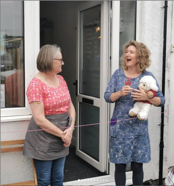 Christine is wearing a blue and white dress and blue leggings and is standing next to Diane who is wearing a pink and orange top, jeans and a grey apron. They are both outside the white front door of the Stitch Station.