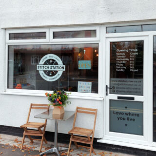 A shop front in a white rendered building. The doors and windows are white UPVC and the shop logo in the window looks like a railway station sign. There are two wooden chairs and a small table with a flower arrangement outside the window.