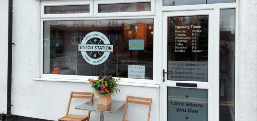 A shop front in a white rendered building. The doors and windows are white UPVC and the shop logo in the window looks like a railway station sign. There are two wooden chairs and a small table with a flower arrangement outside the window.