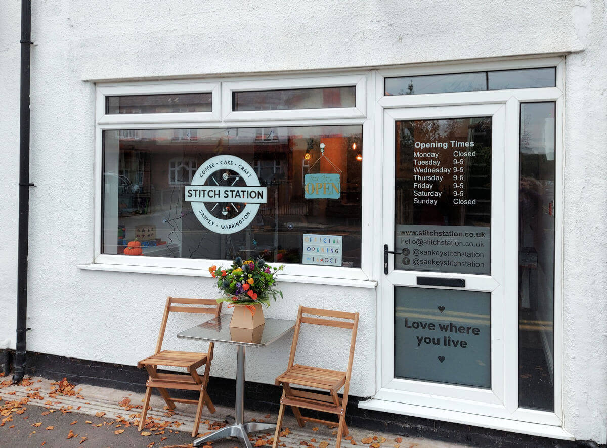 A shop front in a white rendered building. The doors and windows are white UPVC and the shop logo in the window looks like a railway station sign. There are two wooden chairs and a small table with a flower arrangement outside the window.