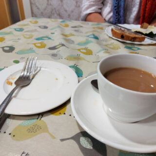 A cup of tea in a saucer is next to a plate which only has crumbs and a fork on it as the cake has all gone. Across the table is another plate with a half-eaten toasted teacake on it.