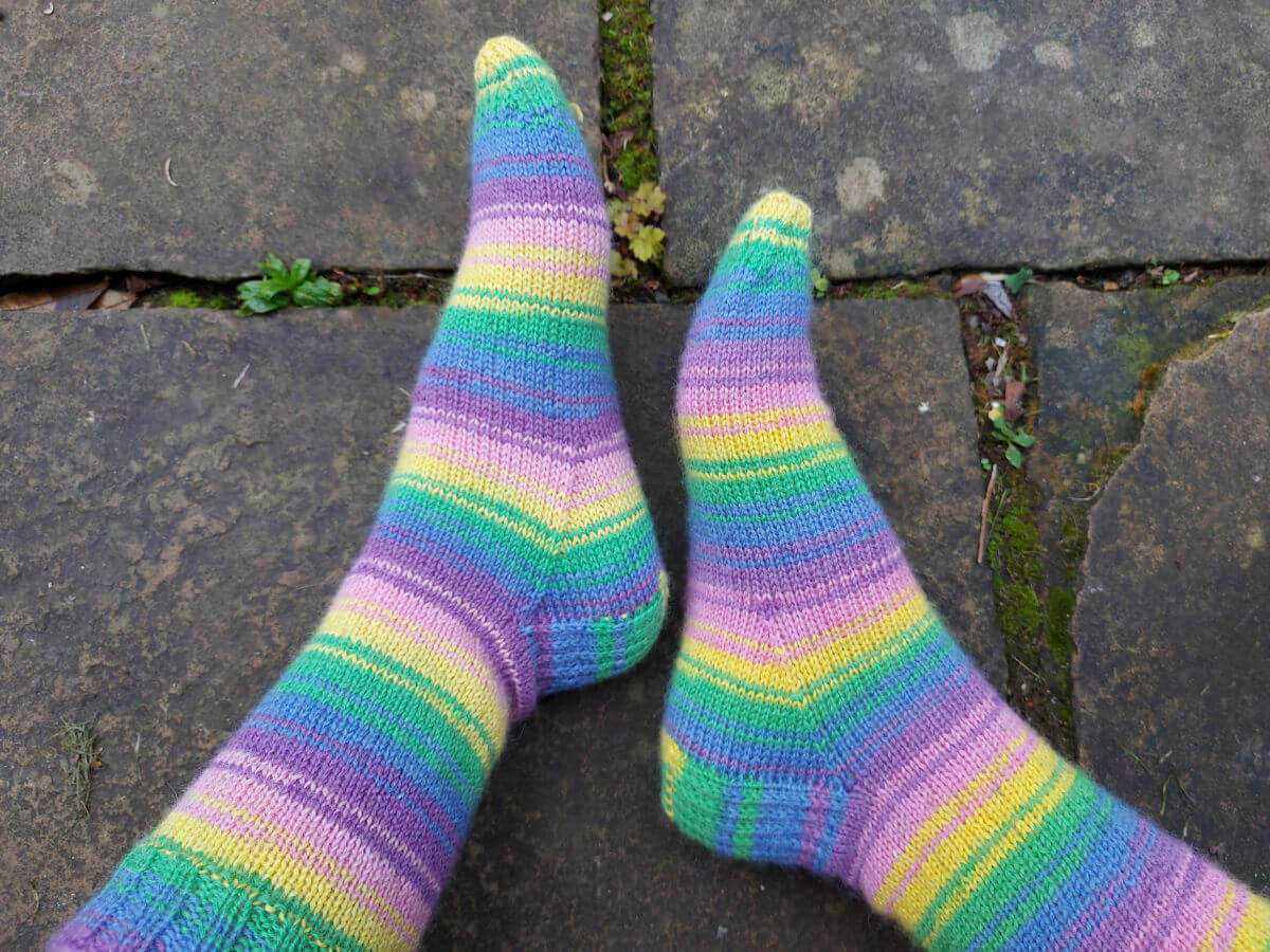 Christine's feet are posed against the grey stone flags so that you can see the stripes of the pink, purple, yellow, blue and green socks.