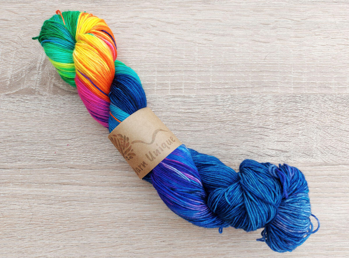 A skein of hand-dyed yarn lying on a beech wooden table. The skein is rainbow-coloured on the left hand side and shades of dark blue on the right.