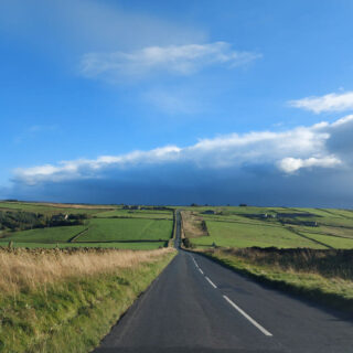 A view from a car looking down a long tarmac road across the moors. The moors to the left and right are green, the sky is bright blue and up ahead are dark rain clouds.