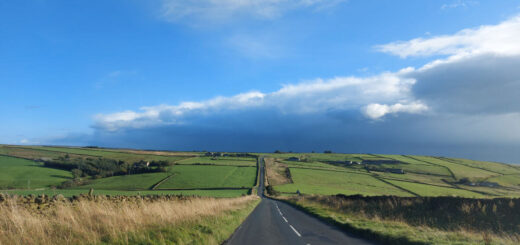 A view from a car looking down a long tarmac road across the moors. The moors to the left and right are green, the sky is bright blue and up ahead are dark rain clouds.